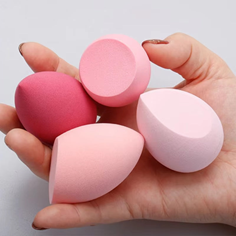 Hand holding four makeup blending sponges in shades of pink and red, including round and teardrop shapes