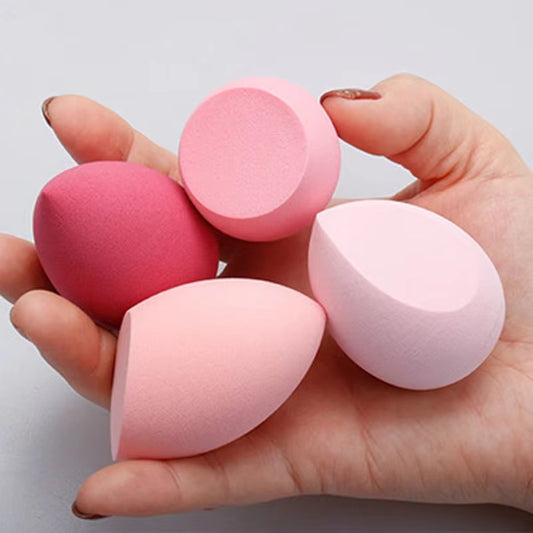 Hand holding four makeup blending sponges in shades of pink and red, including round and teardrop shapes