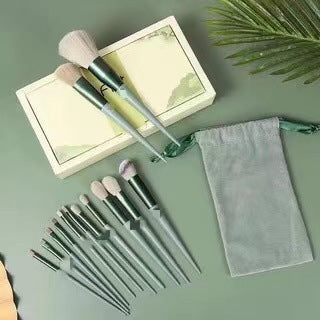 Set of 12 gray makeup brushes with soft bristles, a green drawstring pouch, and a decorative box on a green surface