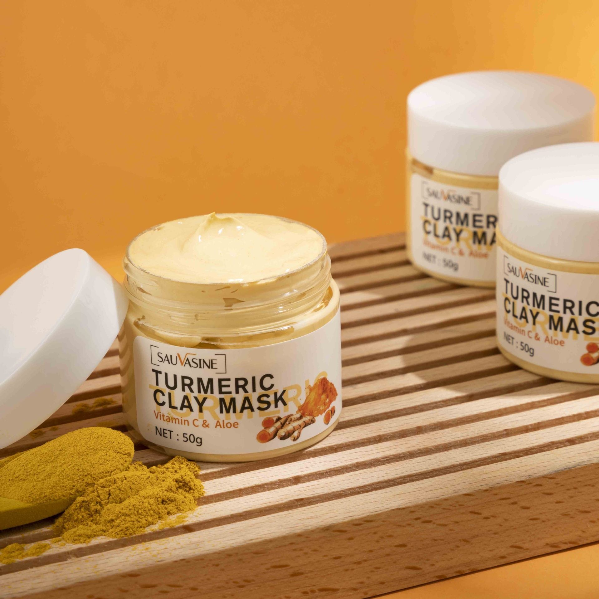 Open jar of turmeric clay mask with Vitamin C & Aloe on wooden tray with turmeric powder and two closed jars
